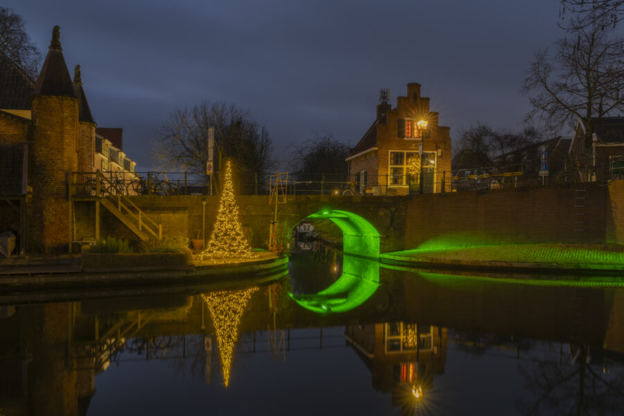 Jsselstein, gelegen in Utrecht (Nederland). Bij deze stadspoort splitst het water zich op. Eén gedeelte verdwijnt onder de brug met het groene licht. Boven de brug: het kleinste restaurant van Nederland. De avond valt, de kerstverlichting gaat aan: een mooie sfeer ontstaat!