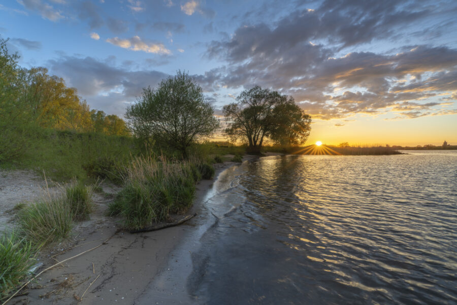 Een mooie zonsondergang aan de rivier nabij Woudrichem (Noord-Brabant, Nederland). Het zonlicht verspreidde haar gouden gloed over de natuur. Golfjes kabbelden aan de oever. Een heerlijk moment zo alleen in dit stukje landschap.