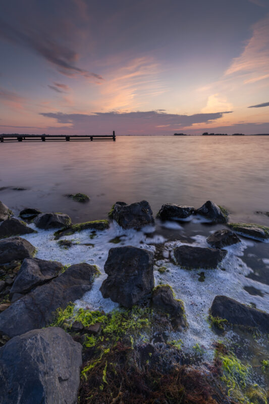 Deze foto is gemaakt tijdens de zonsondergang boven het Grevelingenmeer nabij Bruinisse (Zeeland, Nederland). Mooie warme kleuren sierden de lucht; in combinatie met een kleurrijke voorgrond een fraai geheel. Het schuim tussen de blokken kwam van de redelijk, stevige wind. Al met al een heerlijke avond voor landschapsfotografie