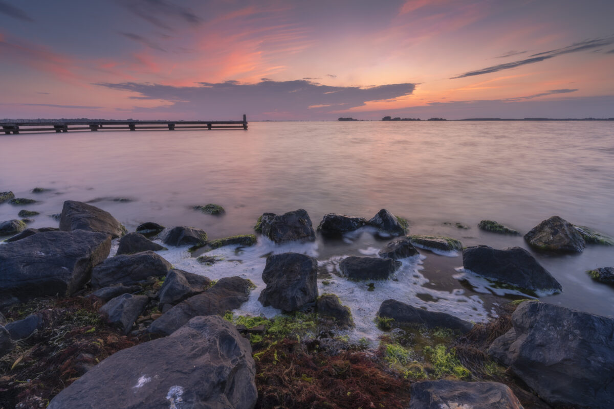 Deze foto is gemaakt tijdens de zonsondergang boven het Grevelingenmeer nabij Bruinisse (Zeeland, Nederland). Mooie warme kleuren sierden de lucht; in combinatie met een kleurrijke voorgrond een fraai geheel. Het schuim tussen de blokken kwam van de redelijk, stevige wind. Al met al een heerlijke avond voor landschapsfotografie